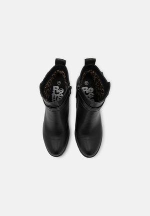 Schwarze Leder-Stiefeletten mit klobigen hohen Absätzen, seitlichen Schnallenriemen und robusten Sohlen, getragen an nackten Beinen vor einem schlichten hellen Hintergrund.