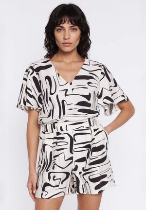 Kobieta stoi ubrana w biały dwuczęściowy strój z czarnymi abstrakcyjnymi wzorami w pociągnięcia pędzla, top z dekoltem w serek i szorty z wysokim stanem.