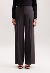 Persona che indossa pantaloni larghi a vita alta di colore grigio scuro, con una maglietta nera a maniche lunghe e sandali neri con le aperture, in piedi su uno sfondo neutro.