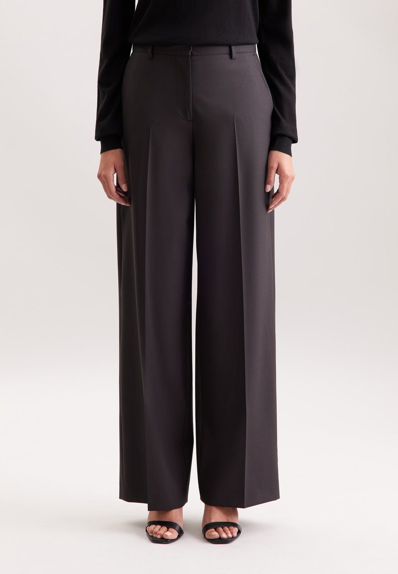 Persona che indossa pantaloni larghi a vita alta di colore grigio scuro, con una maglietta nera a maniche lunghe e sandali neri con le aperture, in piedi su uno sfondo neutro.