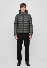 Giacca a vento grigia con zip frontale e cappuccio, caratterizzata da un design trapuntato, materiale leggero e polsini elasticizzati. Abbinata a pantaloni neri.