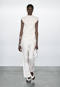 Witte gestructureerde top en brede crèmekleurige broek; model houdt een geweven tas vast en draagt witte sandaaltjes met bandjes; zilveren manchette en oorbellen accentueren de look.