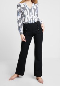 Pantaloni neri a vita alta con dettagli a bottoni, abbinati a una blusa a fantasia floreale e a righe con scollo a V e maniche lunghe.