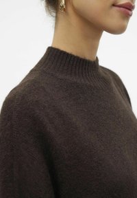 Pull en maille marron à col haut avec une finition texturée. Tissu doux et détail encolure côtelée. Boucle d'oreille dorée visible, mettant en valeur le décolleté.