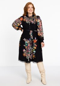 Schwarzes Kleid mit lebhaftem floralen Stickereien und transparenter Ärmeln, tailliert mit einem schwarzen Gürtel. Kombiniert mit beigen kniehohen Stiefeln.