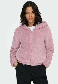 Roze nepbonten zip-up hoodie met capuchon en afgeronde zoom. Voorzien van zijzakken en geribbelde manchetten. Draagt over een wit shirt.