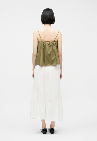 Γυναίκα με κοντά μαύρα μαλλιά που φοράει ελιάς πράσινο αμάνικο τοπ και μακριά λευκή φούστα, γυρισμένη προς τα πίσω σε απλό λευκό φόντο.