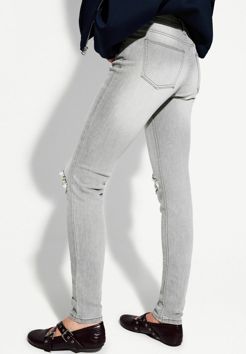 Jean skinny gris avec un genou déchiré, design à cinq poches et tissu légèrement décoloré, associé à des chaussures plates sombres.
