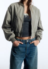 Cazadora bomber recortada de algodón verde oliva con puños acanalados y cremallera frontal, combinada con jeans de mezclilla azul de talle alto.