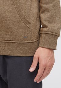 Bruine getextureerde sweatshirt met een kangoeroezak en geribde manchetten, voorzien van een klein logo label nabij de zoom. Close-up van de mouw en hand.