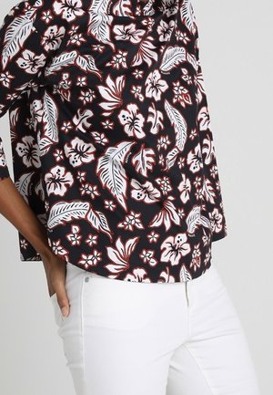 Sort skjorte med hvidt og rødt tropisk blomsterprint båret med hvide bukser, hånd hvilende på hoften.