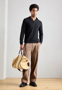 Zwarte gebreide trui met een brede kraag, bruine wijde broek, en een grote gele sporttas met donkerbruine accenten. Zwarte schoenen.