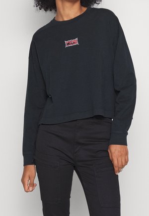 Person trägt schwarzes, langärmeliges, kurz geschnittenes Shirt mit kleinem rotem Logo auf der Brust und schwarze Hose mit Cargotaschen, steht vor einem schlichten Hintergrund.