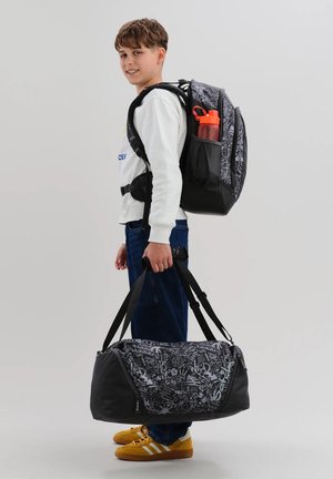 Junge mit kurzem braunem Haar, lächelnd, trägt weißen Pullover und Jeans, trägt gemusterten schwarzen Rucksack mit orangefarbener Wasserflasche und passender Sporttasche.