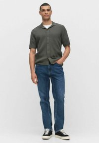 Olijfgroene shirt met korte mouwen en een gestructureerd patroon, knoopsluiting, gecombineerd met blauwe jeans en zwarte sneakers.