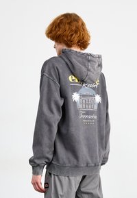 Sudadera con capucha gris con un diseño gráfico en la parte trasera que representa palmeras y un club de playa, confeccionada con tela suave y cómoda.