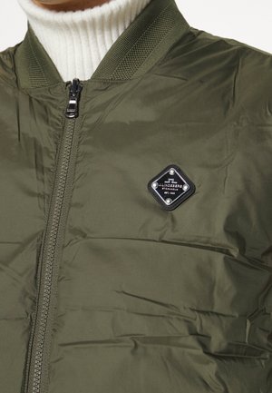 Gros plan d'une veste zippée verte avec col côtelé et écusson de marque noir en forme de losange sur la poitrine, portée sur un pull à col roulé blanc.