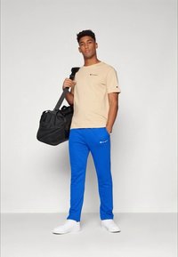 Beige t-shirt och blåa sweatpants med logodetaljer. Modellen håller en svart duffelväska och har på sig vita sneakers. En enkel bakgrund.