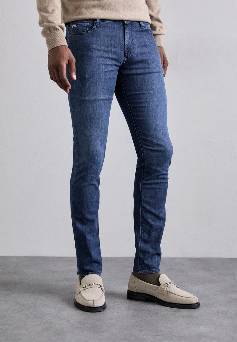 Blugi slim-fit de culoare albastru închis, cu o textură netedă, bandă de talie sigură și un design cu cinci buzunare, asortați cu mocasini de culoare bej deschis.