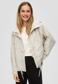 Lichtbeige jas met een capuchon, voorrits, twee borstzakken en verstelbare trekkoorden aan de onderkant. Draag het over een witte top.