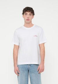 Λευκό βαμβακερό μπλουζάκι με κοντά μανίκια, στρογγυλή λαιμόκοψη και κόκκινο κεντητό λογότυπο στο αριστερό στήθος. Το μοντέλο φοράει ανοιχτό μπλε τζιν παντελόνι.