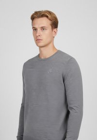 Ung man med blont hår som bär en enkel grå tröja med långa ärmar, stående mot en ljusgrå bakgrund och vänd mot kameran.