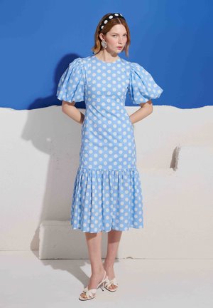 Femme portant une robe midi bleu clair à pois blancs, manches bouffantes, ourlet à volants, debout devant un mur bleu et blanc.