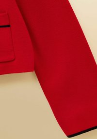 Punainen pitkähihainen neulepaita taskulla, jossa ohut musta raita beigen taustalla, näyttää kangasrakenteen ja hihansuun yksityiskohdan.