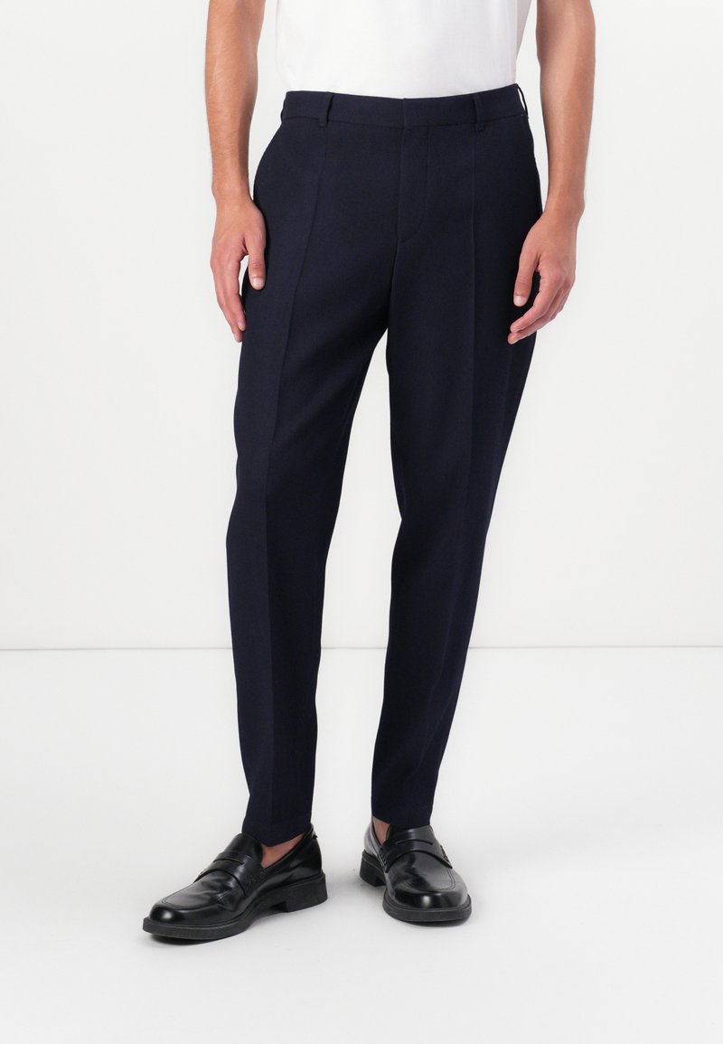 Sötétkék szabott nadrág sima anyagból, slim fit fazonnal, éles hajtással és övhurkokkal, fekete bőr cipőkkel párosítva.