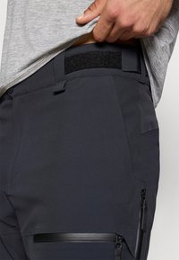 Σκούρο γκρι παντελόνι με ζώνη μέσης με βέλκρο, τσέπη με πλευρικό φερμουάρ και λεία υφή υφάσματος. Ο σχεδιασμός περιλαμβάνει διακριτικές λεπτομέρειες κοντά στη μέση.