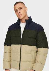 Padded bunda s vysokým golierom, so zafarbenými blokmi námorníckej modrej, tmavozelenej a béžovej farby. Obsahuje predný zips a bočné vrecká.