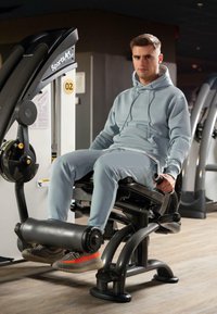 Attrezzatura da palestra con un design nero e argento. Una persona con un hoodie azzurro chiaro e pantaloni utilizza una macchina per l'estensione delle gambe con supporto per i piedi imbottito.