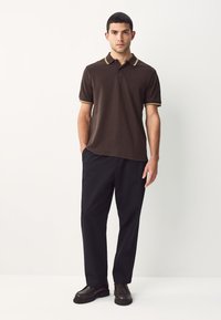 Brun polotrøje med gule og hvide accentstriber på kraven og ærmerne, kombineret med sorte bukser og mørke sko.