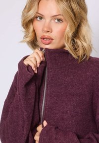 Burgunderfarbene Fleecejacke mit Reißverschluss, hohem Kragen, weicher Textur und langen Ärmeln. Auffälliges silbernes Reißverschlussdetail an der Vorderseite.