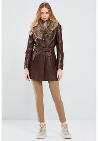 Brown leather coat with a fur collar, belted waist, and two front pockets. The model wears tan leggings and beige high-top sneakers.