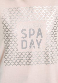 T-shirt di un delicato rosa pallido con un motivo a scallop in argento metallico e testo che dice "Storie di una giornata alla spa in montagna" in caratteri grassetto e corsivo.