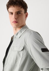 Ανοιχτό πράσινο καπιτονέ μπουφάν με φερμουάρ μπροστά, αναδιπλούμενο γιακά και τσέπη στο στήθος. Διαθέτει λογότυπο στην μανσέτα. Υφασμάτινη επιφάνεια.