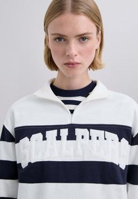 Jersey con rayas blancas y azul marino y cuello con cremallera, con la palabra "SCALPERS" en letras en relieve en el pecho, confeccionado en una tela suave.