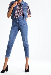 Denim vest, geruit shirt en high-waisted blauwe spijkerbroeken met opgerolde boorden. Zwarte hakken maken de outfit af. Heldere, gladde texturen en klassieke snits.