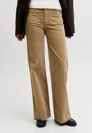 Personne portant un pantalon large taille haute marron clair, des chaussures noires, un haut blanc et une veste rayée foncée, debout sur un fond uni.