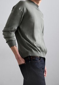 Grön stickad bomullströja med rund halsringning, åtsittande muddar och avslappnad passform, kombinerad med mörka denimjeans med kontrasterande sömmar.