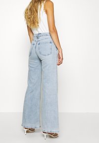 Light blue high-waisted wide-leg jeans made of denim, featuring two back pockets and subtle fading, paired with white open-toed heels.