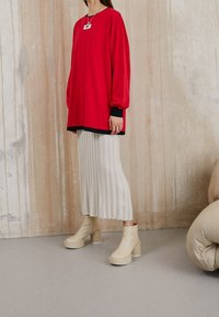 Kvinna som bär en röd oversized topp, en veckad vit kjol, beige plattform ankelstövlar och ett hängande halsband mot en strukturerad vägg.