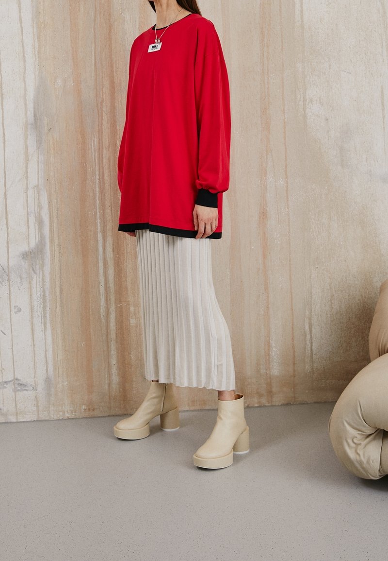 Kvinna som bär en röd oversized topp, en veckad vit kjol, beige plattform ankelstövlar och ett hängande halsband mot en strukturerad vägg.