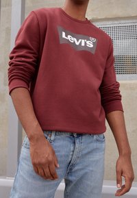 Sudadera color burdeos con cuello redondo, que presenta un gran logo blanco de Levi's. Material de algodón, mangas ajustadas, combinada con jeans de color azul claro.