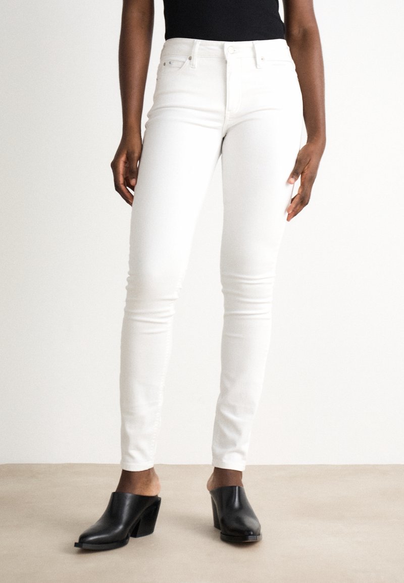 Jeans bianchi a taglio slim con una texture liscia e vita alta. Abbinati a mule neri open-toe, che mostrano un design con tacco a blocco.