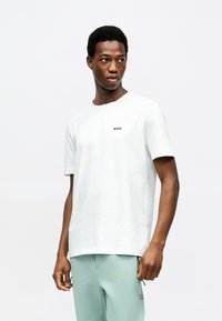 Νεαρός άνδρας που φοράει απλό λευκό T-shirt BOSS και ανοιχτό πράσινο παντελόνι, στέκεται μπροστά σε λευκό φόντο, κοιτάζοντας προς τα αριστερά του.