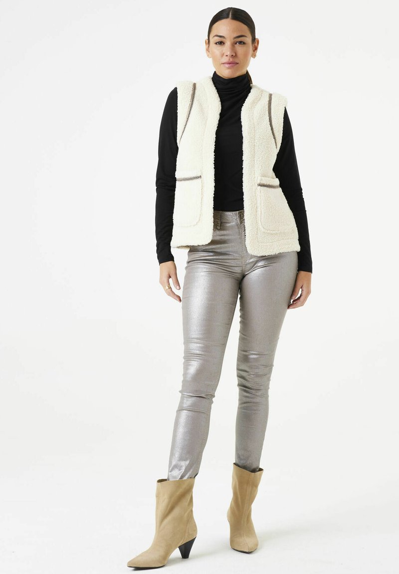 Crème nep bont vest met ritszakken, gedragen over een zwarte coltrui. Zilveren glanzende broek en beige enkellaarsjes maken de outfit compleet.