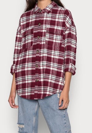 Persona che indossa una camicia a quadri bordeaux e bianca a maniche lunghe con polsini arrotolati e jeans azzurri strappati.
