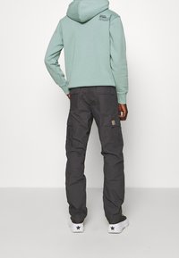 Hellgrüner Hoodie mit großer Kapuze und grafischem Druck auf dem Rücken, kombiniert mit dunkelgrauen Cargo-Hosen mit Seitentaschen und einem Patch.